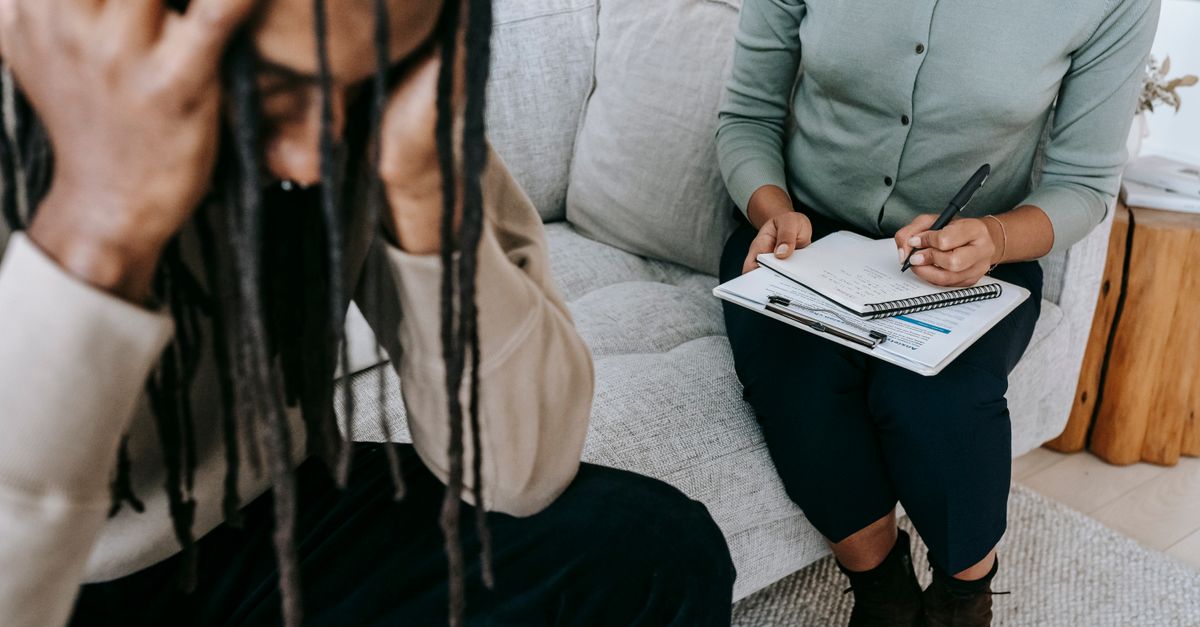 Crop concerned African American male sitting on sofa with female psychologist and holding head in hands in dismay during psychotherapy session