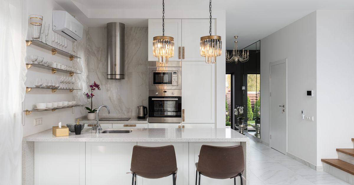 Interior of contemporary kitchen with modern appliances and chairs at white counter near  wall with shelves in spacious apartment near stairs