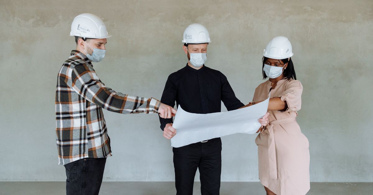 Three architects wear masks and helmets while reviewing construction plans indoors.