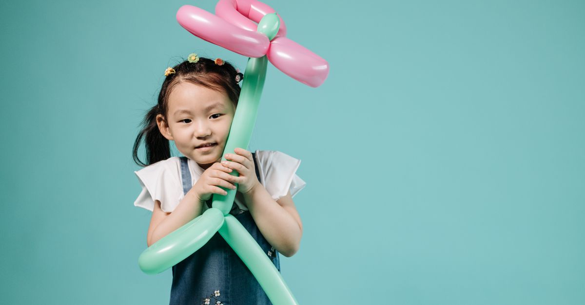 Adorable Asian girl in denim holding a pink and green balloon shaped like a flower against a teal backdrop.