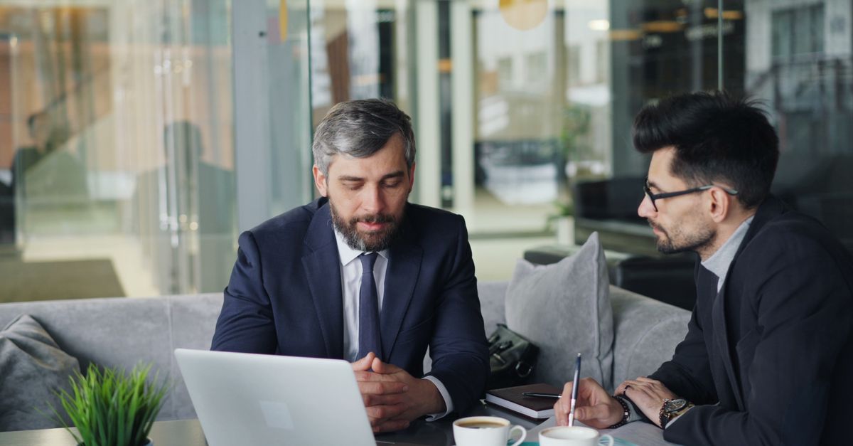 Two businessmen in suits discussing work at a cafe with laptops and coffee.