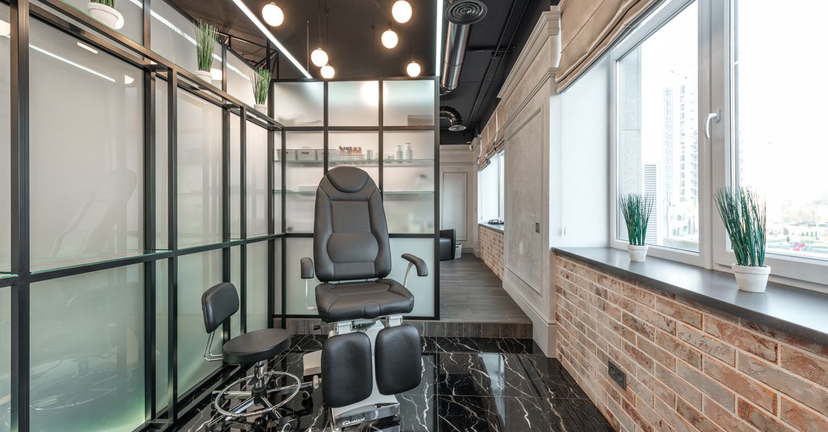 Black leather pedicure chair placed between glass door and window in modern beauty salon with decorated plants and glowing lamps