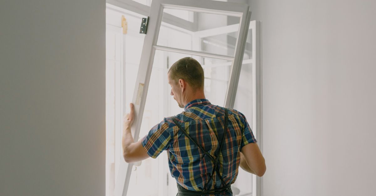 Back view of anonymous male fitter in checkered shirt and apron mounting window with hinges near white walls at home