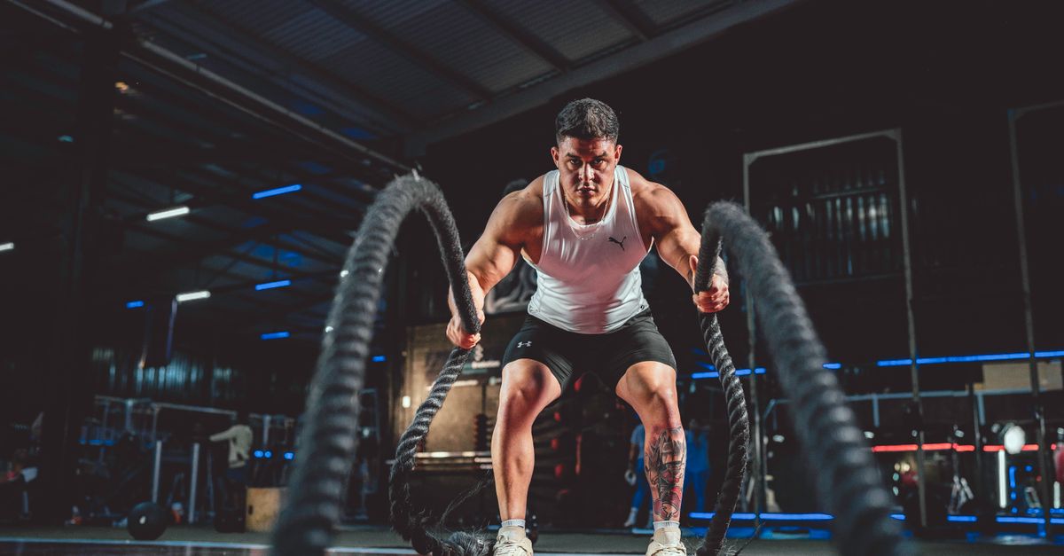 Focused man in white tank top performing battle rope exercise in a modern gym.