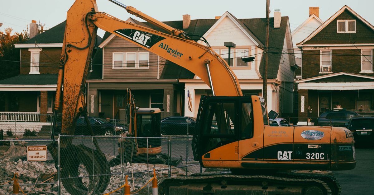 Excavator working in a suburban neighborhood construction site, with residential houses in the background.
