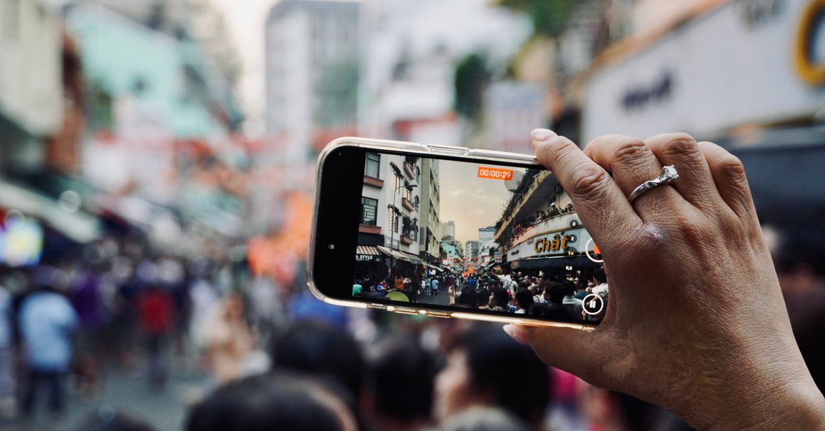 Hand holding smartphone capturing bustling city street scene with crowd.