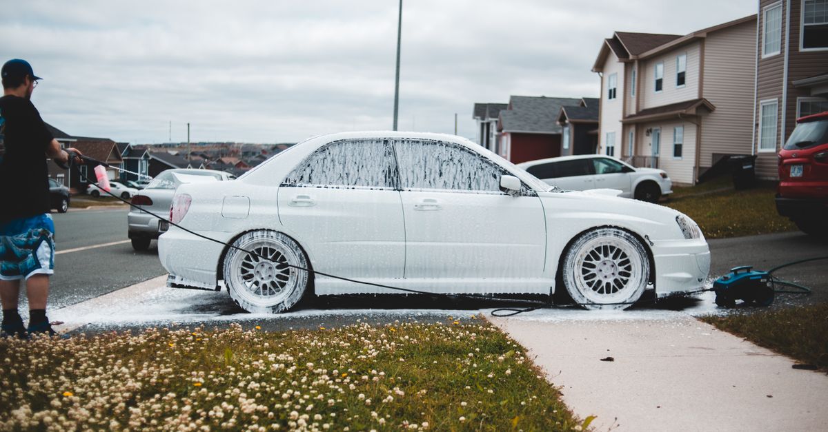 Foamed car on driveway and sidewalk being washed by male driver at daytime