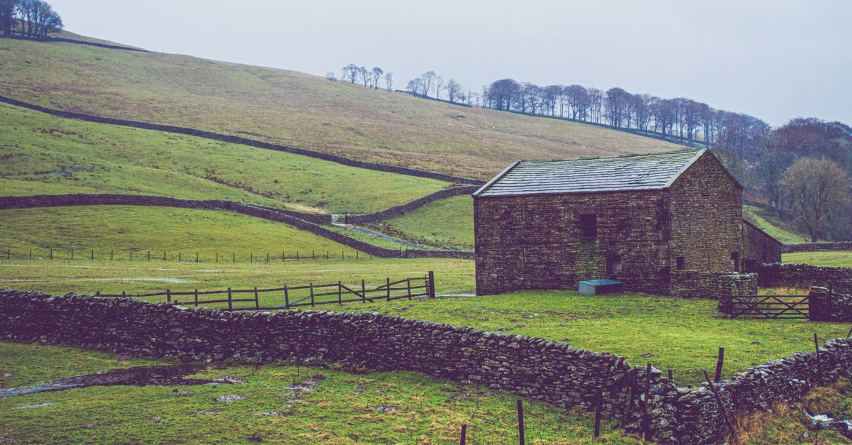 A solitary stone barn on a grassy hillside with surrounding dry stone walls on a cloudy day.