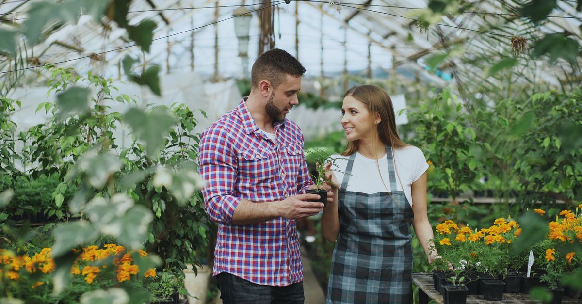 Smiling young couple tending plants inside a vibrant greenhouse filled with flowers.