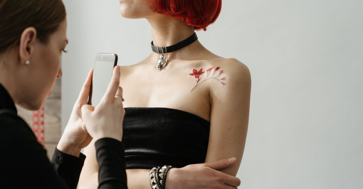 A woman with red hair and a shoulder tattoo being photographed by another woman in a tattoo studio.