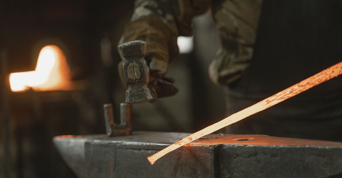 A blacksmith uses a hammer to shape hot iron on an anvil in a dimly lit workshop.