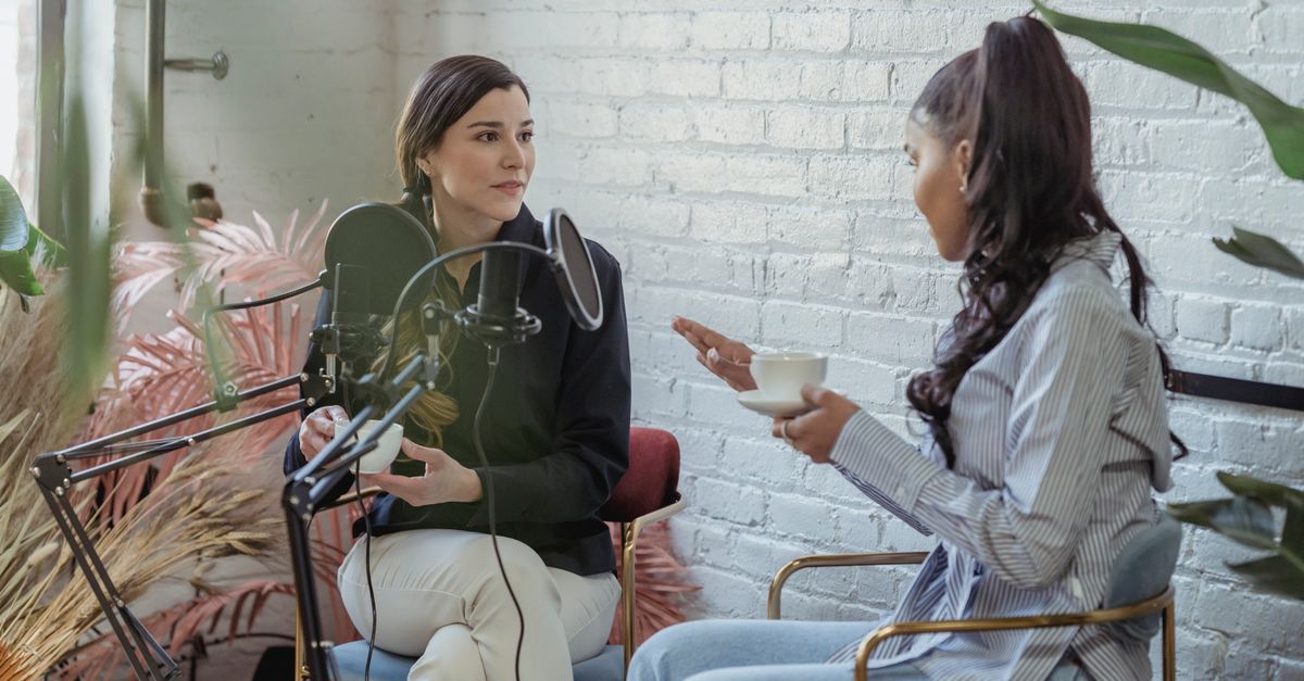 Serious host listening to African American female guest giving interview while sitting with cup of coffee near mic