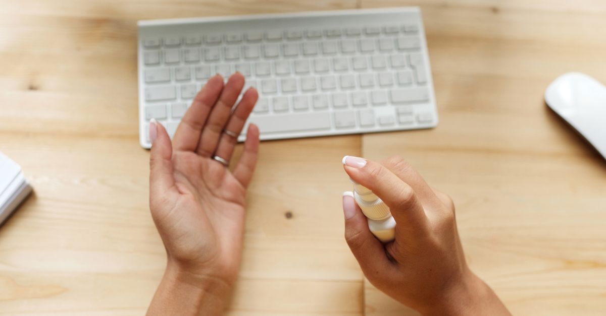 Close-up of hands using sanitizer spray near a keyboard on a wooden desk.