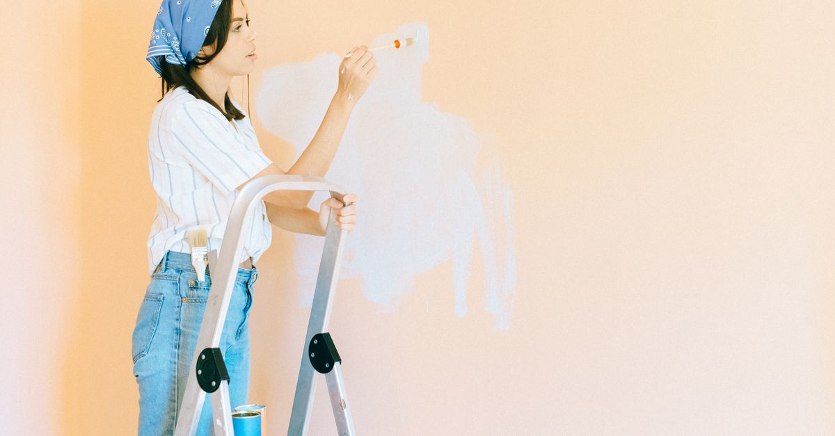 Young woman in bandana painting wall with white paint using a brush, standing on a ladder indoors.