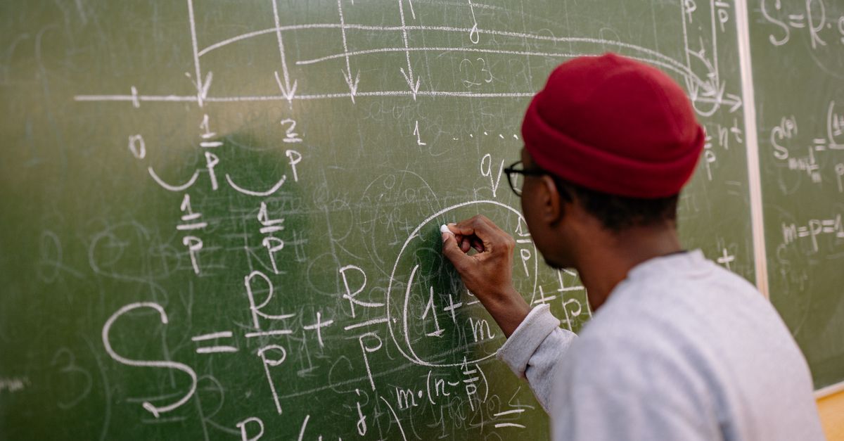 Student writing complex mathematical equations on a chalkboard in a classroom setting.