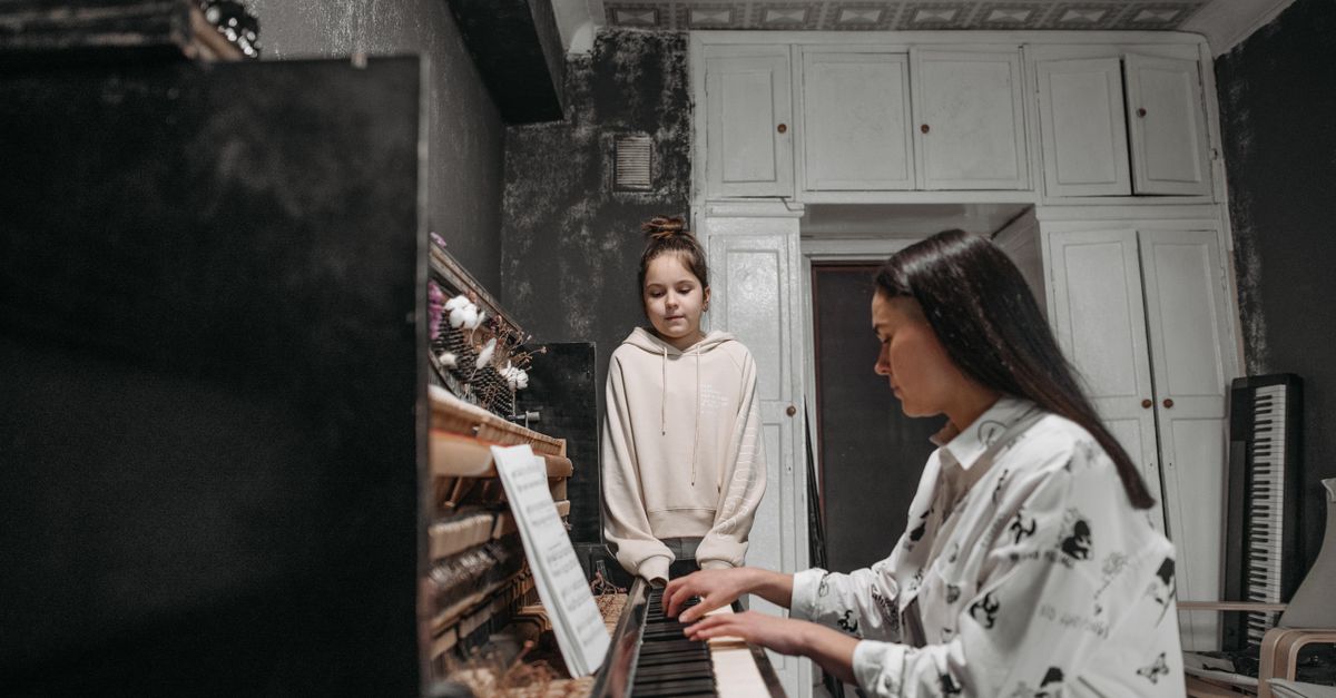 A student observes her teacher practicing piano in a cozy home music studio setting.