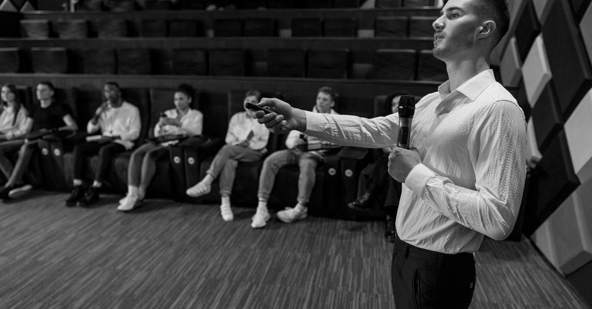 Monochromatic image of a speaker presenting to a small audience in a stylish auditorium.