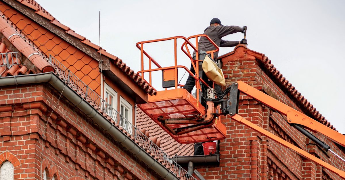 Roofer in a cherry picker conducting maintenance on a red brick building with a tiled roof.
