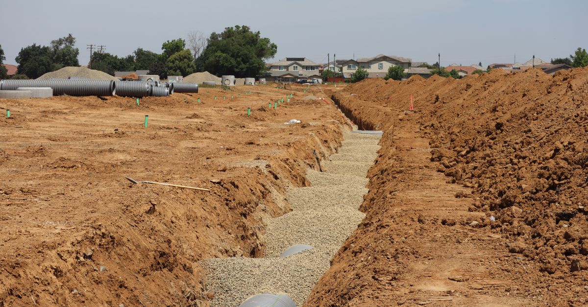Wide view of a construction site with trenches and large pipes in a suburban setting under a clear sky.