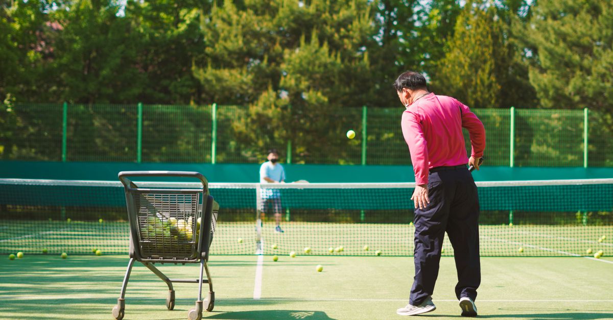 Two men practicing tennis on a sunny outdoor court with a ball cart.