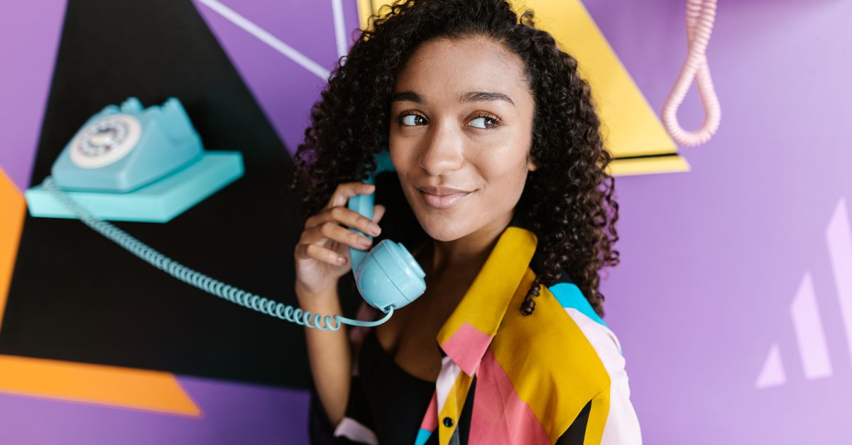 A woman with curly hair smiles while using a vintage blue telephone against a colorful backdrop.