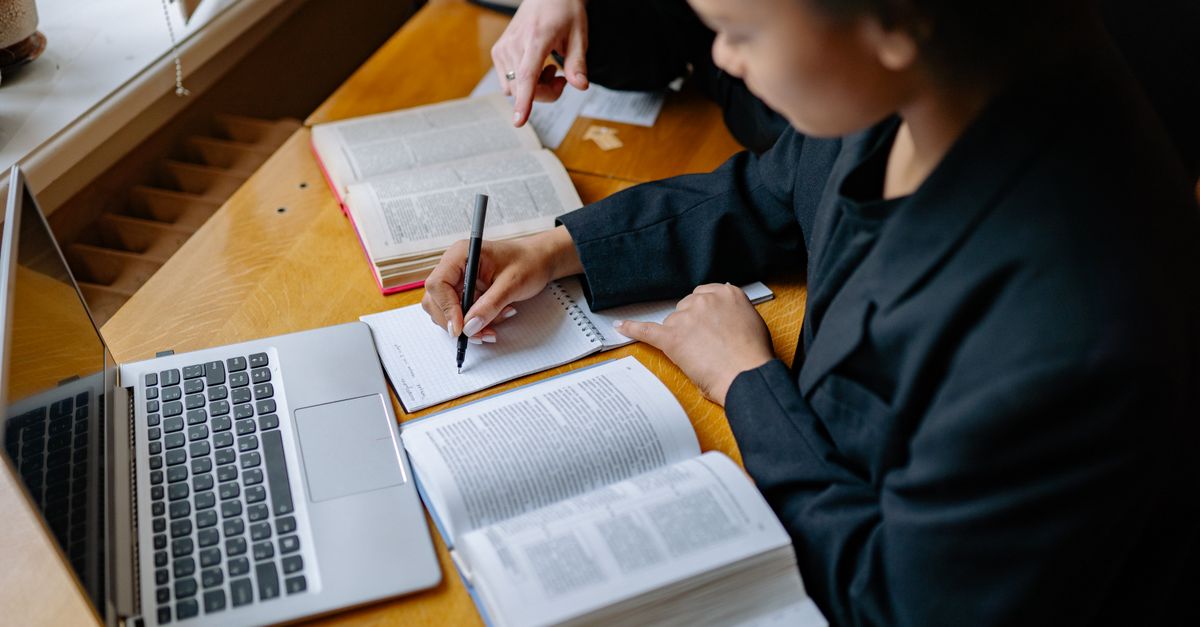 Two students engaged in study, taking notes with books and a laptop on a wooden desk indoors.