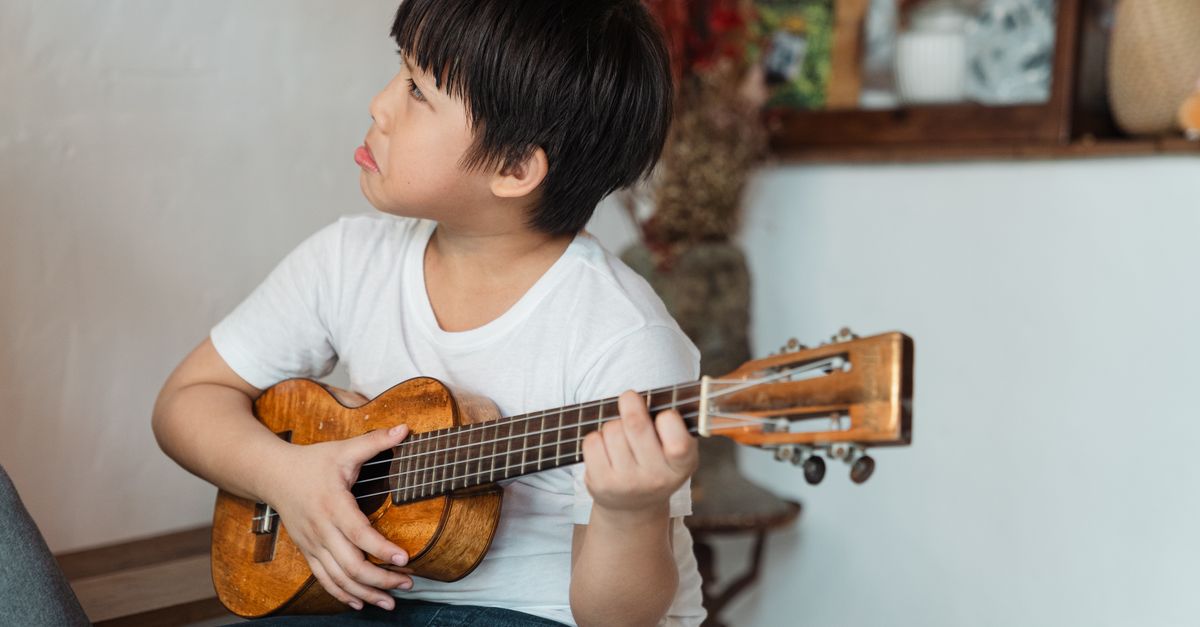 Melancholic ethnic boy in casual wear playing classic musical instrument while sitting near kitchen and looking away at home on blurred background