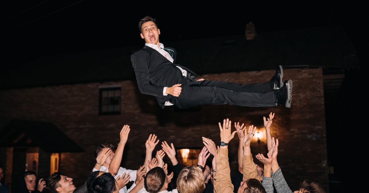 A lively wedding party scene with guests lifting a man in the air during the night celebration.