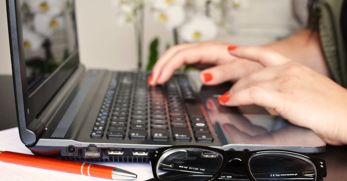 Close-up of a woman's hands typing on a laptop with glasses and a notepad on the desk.